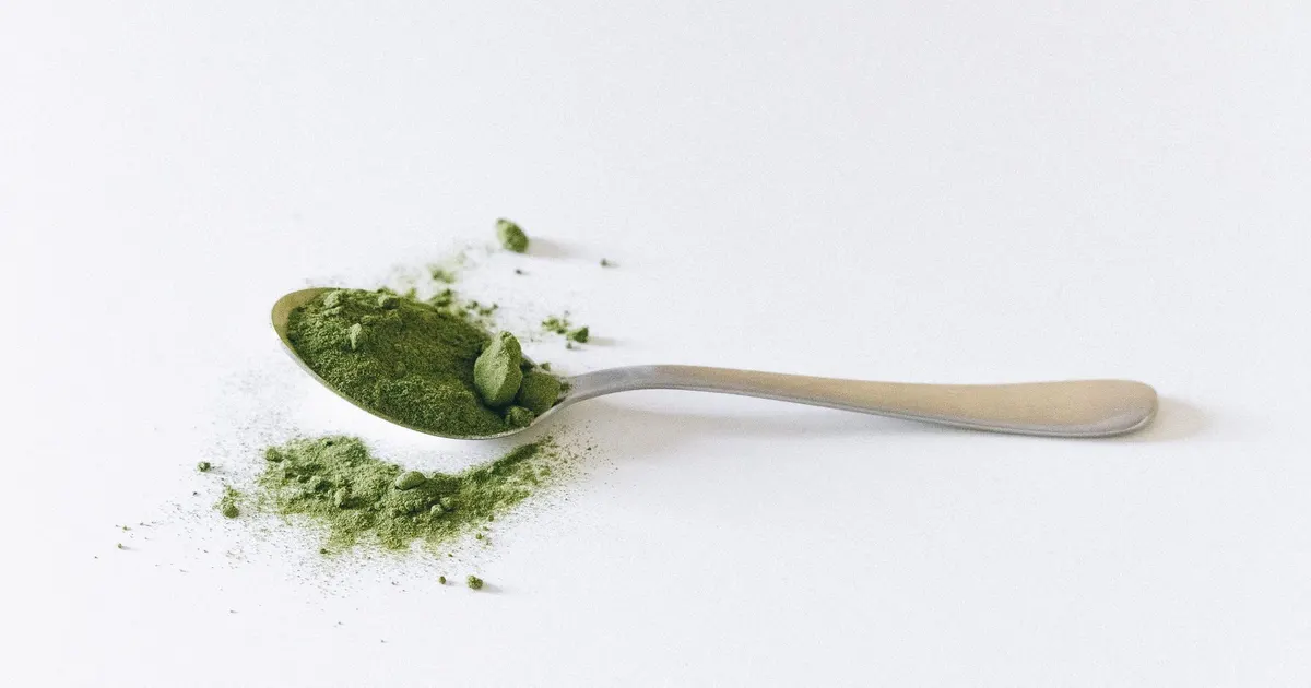A metal spoon of bright green tea powder with loose powder on a white surface, the ingredient bought as green tea powder in bulk